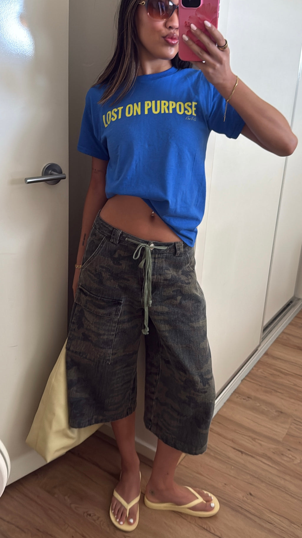 A woman takes a mirror selfie wearing a bright blue t-shirt with yellow text that reads 'LOST ON PURPOSE', paired with dark gray camouflage wide-leg cargo shorts and beige flip-flop sandals.